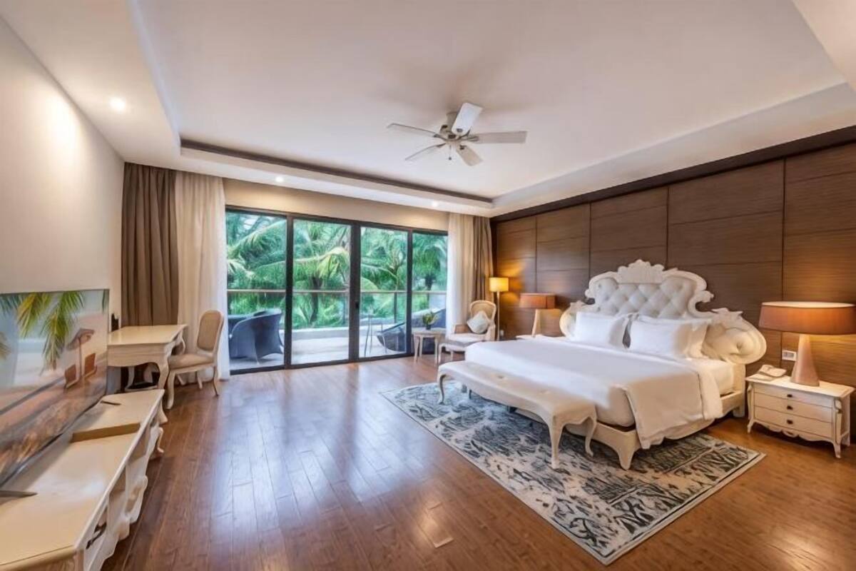 A spacious bedroom with a large bed, a desk, and a chair, a ceiling fan, and a view of greenery through the window.