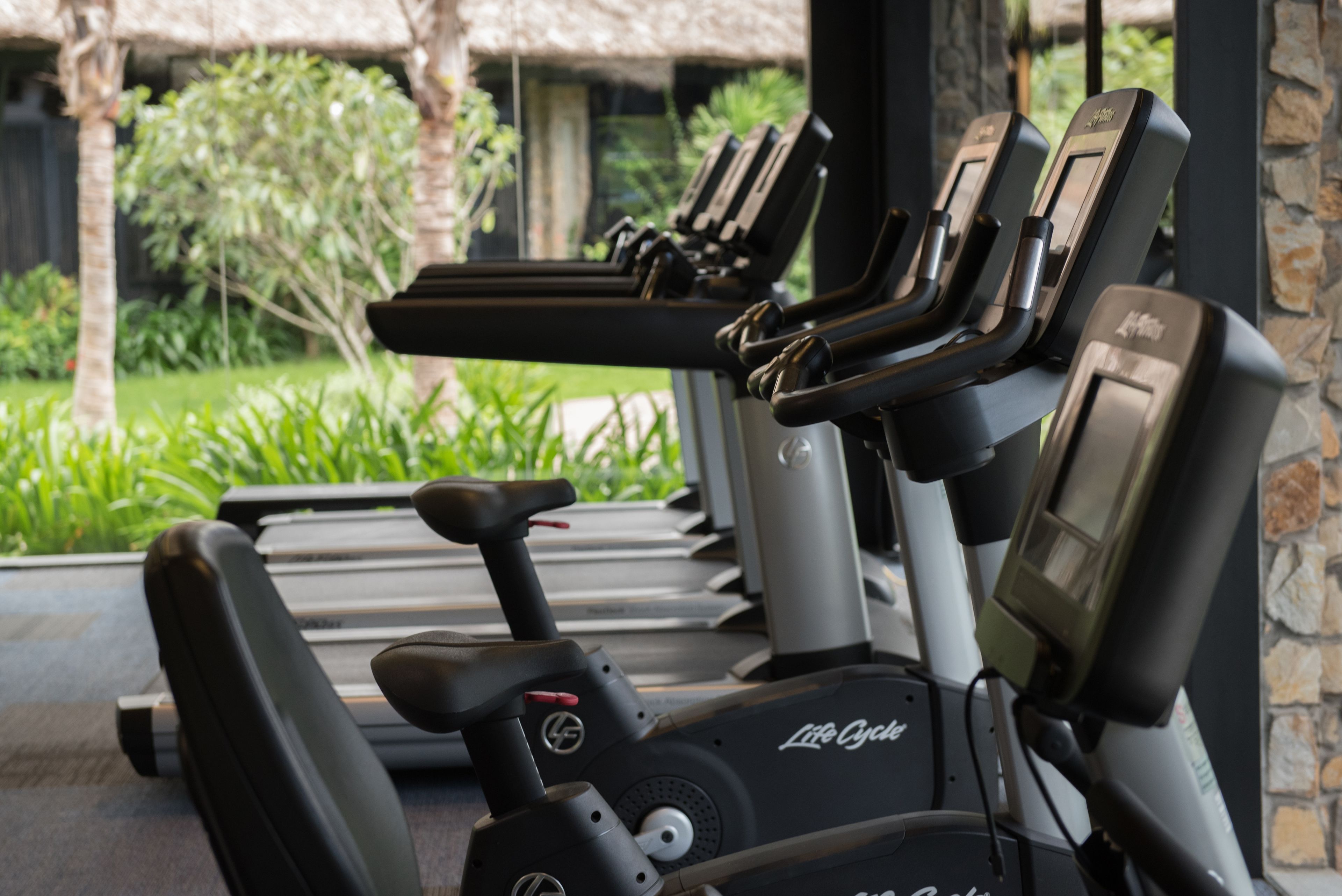 A row of stationary bikes labeled "Life Cycle" in a covered area with a view of greenery and a stone wall.