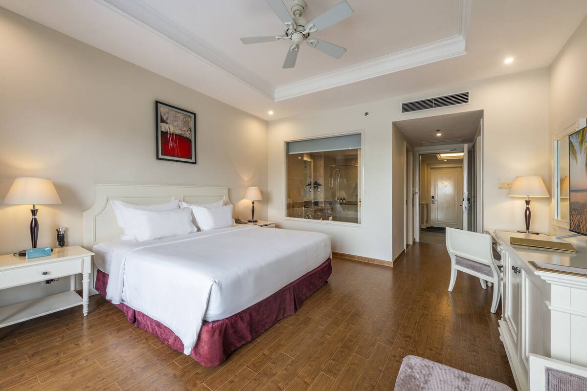 A hotel room with a large bed, bedside tables, a desk, and a chair. There is a ceiling fan, a window, and a door leading to another room.