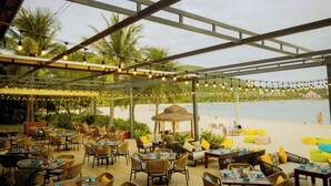 An outdoor dining area with tables and chairs, a thatched-roof structure, and a view of the beach and sea. - Vinpearl Resort & Spa Nha Trang Bay (Nha Trang)