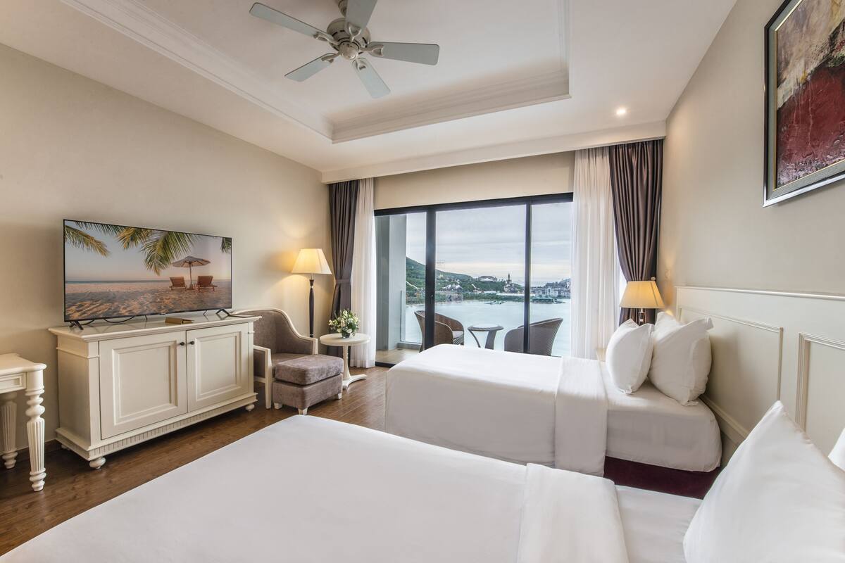 A hotel room with two beds, a TV, a desk, and a view of a cityscape.