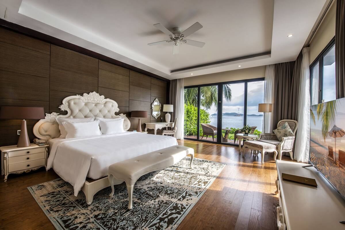 A spacious bedroom with a large bed, a ceiling fan, a view of the outdoors, and a balcony with seating.