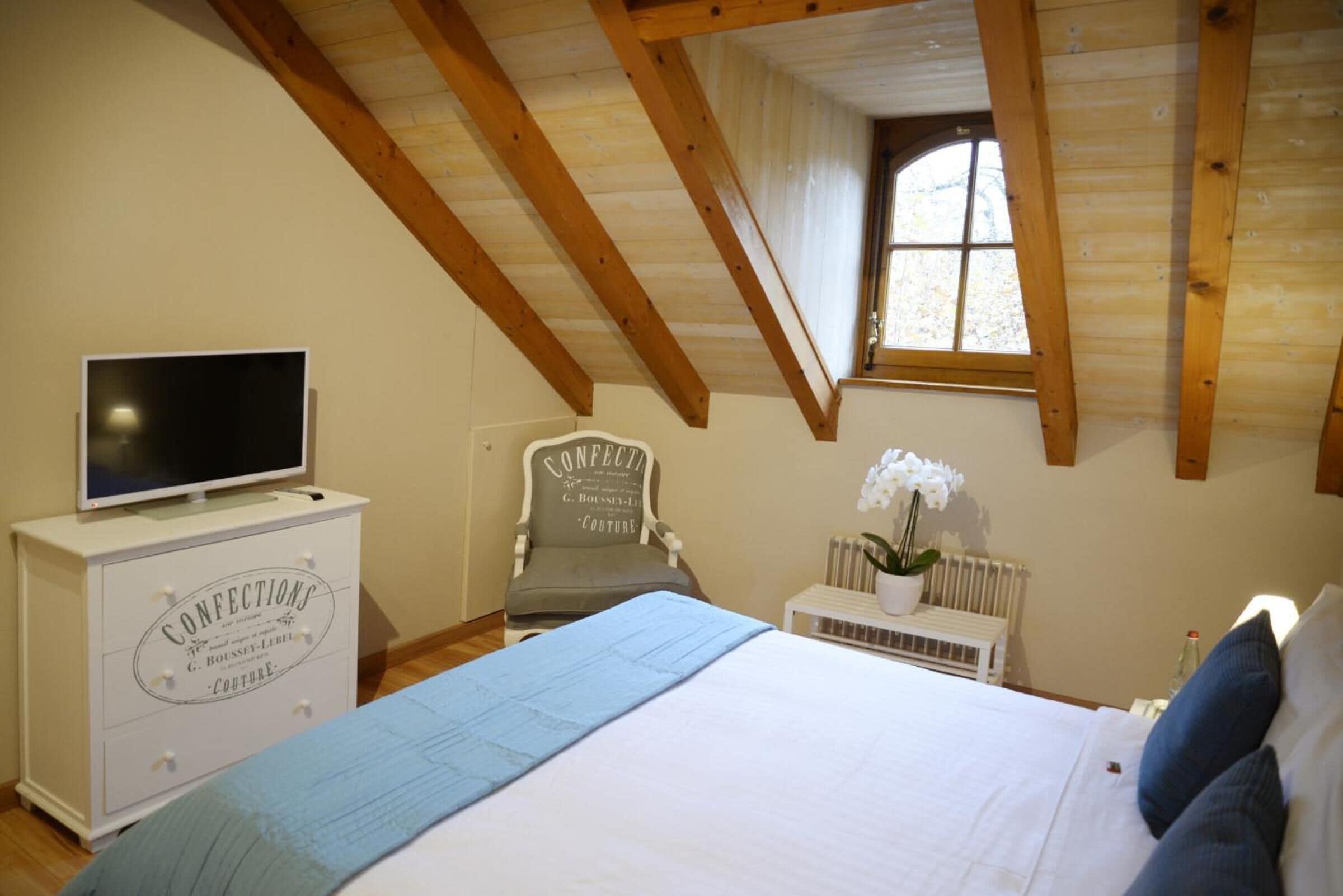 A bedroom with a bed, a dresser, a television, a chair, a window, and a radiator.