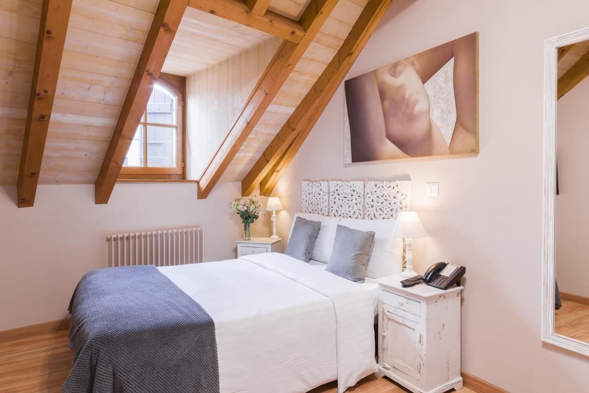 Quarto com teto inclinado, cama com cobertor cinzento, luminárias de cabeceira, mesa de cabeceira com telefone e um espelho grande.