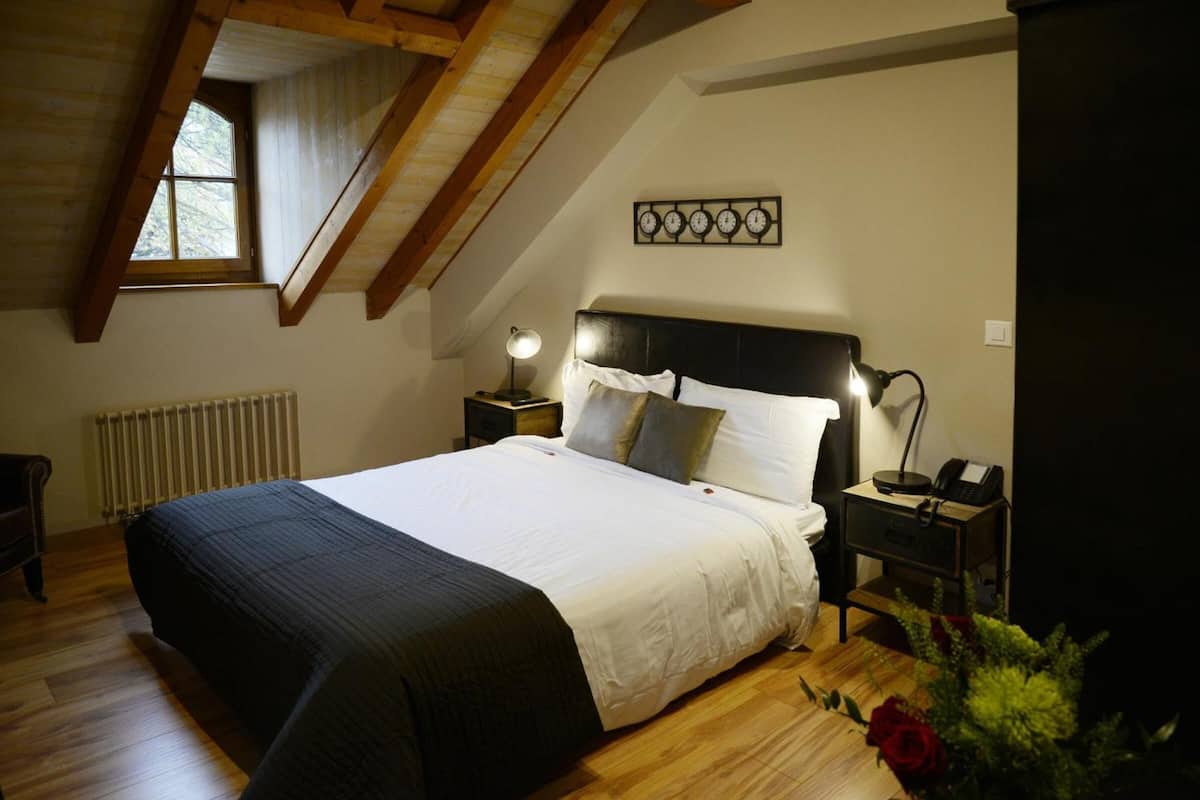 Quarto com uma cama grande, vigas de madeira, radiador, relógio e mesa de cabeceira com luminária.