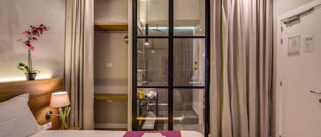 A hotel room with a bed, a nightstand with a lamp, a potted plant, and a bathroom visible through a glass door.