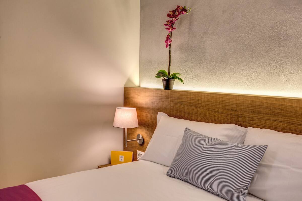 A neatly made bed with white and striped pillows, a bedside table with a lamp, a small potted plant, and a yellow item on the table.