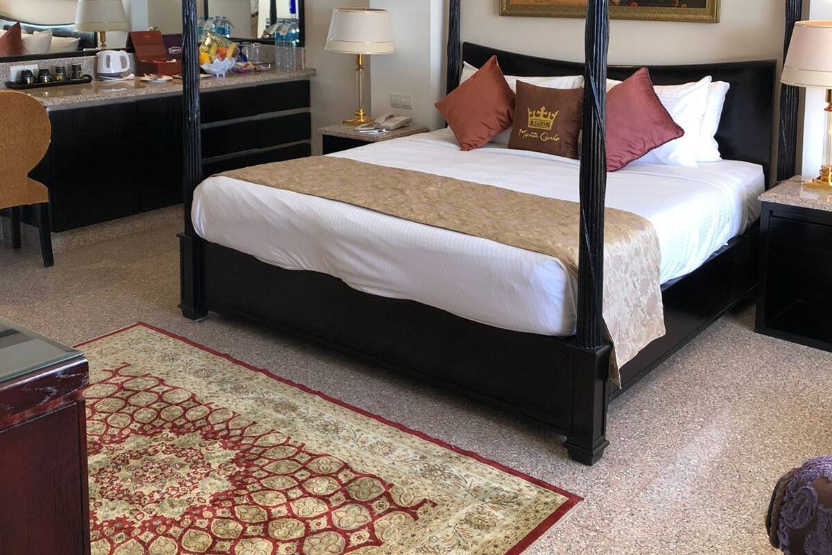 A four-poster bed with a red and beige bedspread, a nightstand with a lamp, a desk with a mirror, and a large painting on the wall.