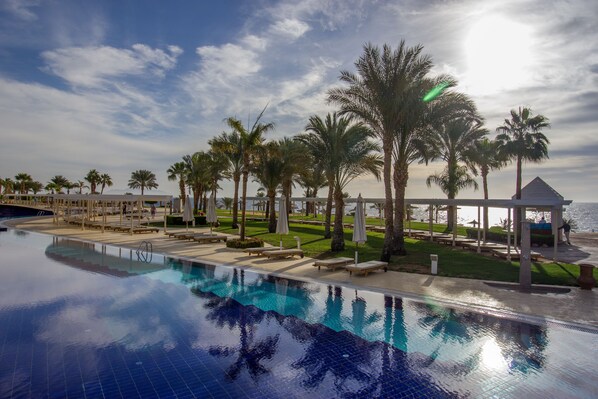 Un área junto a la piscina con palmeras, sillas de descanso y un pabellón con vistas al mar.