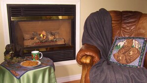 Una sala de estar acogedora con chimenea, una mesa verde con galletas y una taza, y un sillón de cuero con un cojín con motivos de oso.