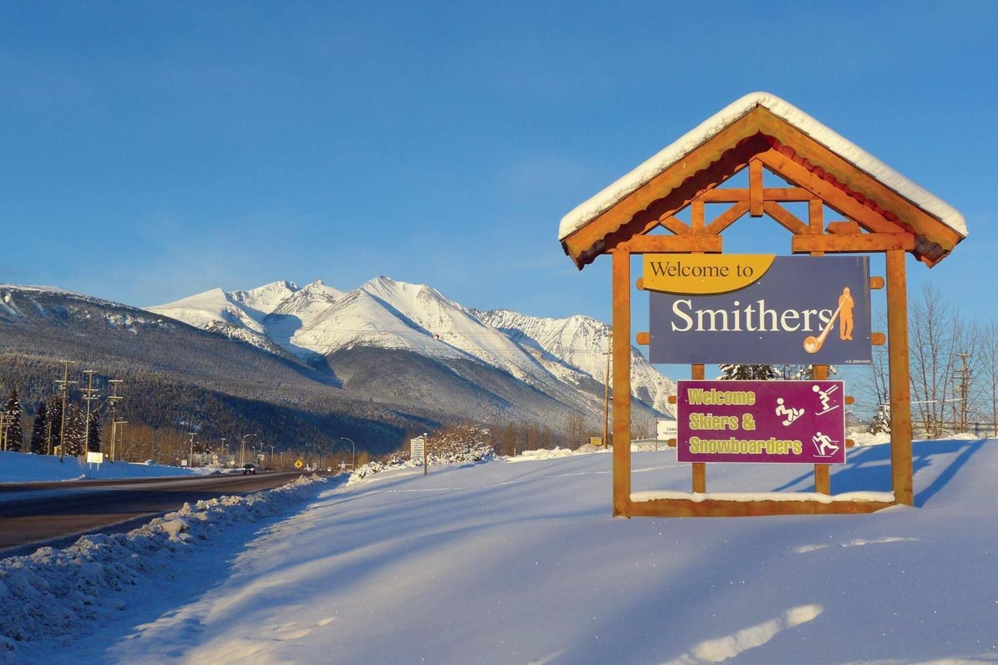 A snow-covered road with a sign welcoming visitors to Smithers, indicating facilities for skiers and snowboarders.