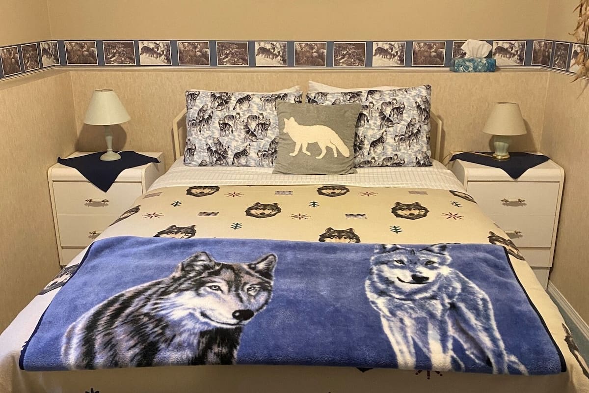 A bedroom with a bed featuring wolf-themed bedding, two bedside tables with lamps, and a framed picture of a whale on the wall.