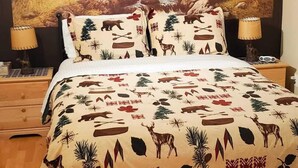 Una cama con cubrecama de motivos forestales, una mesita de noche con lámpara y un tapiz mural con paisaje montañoso.