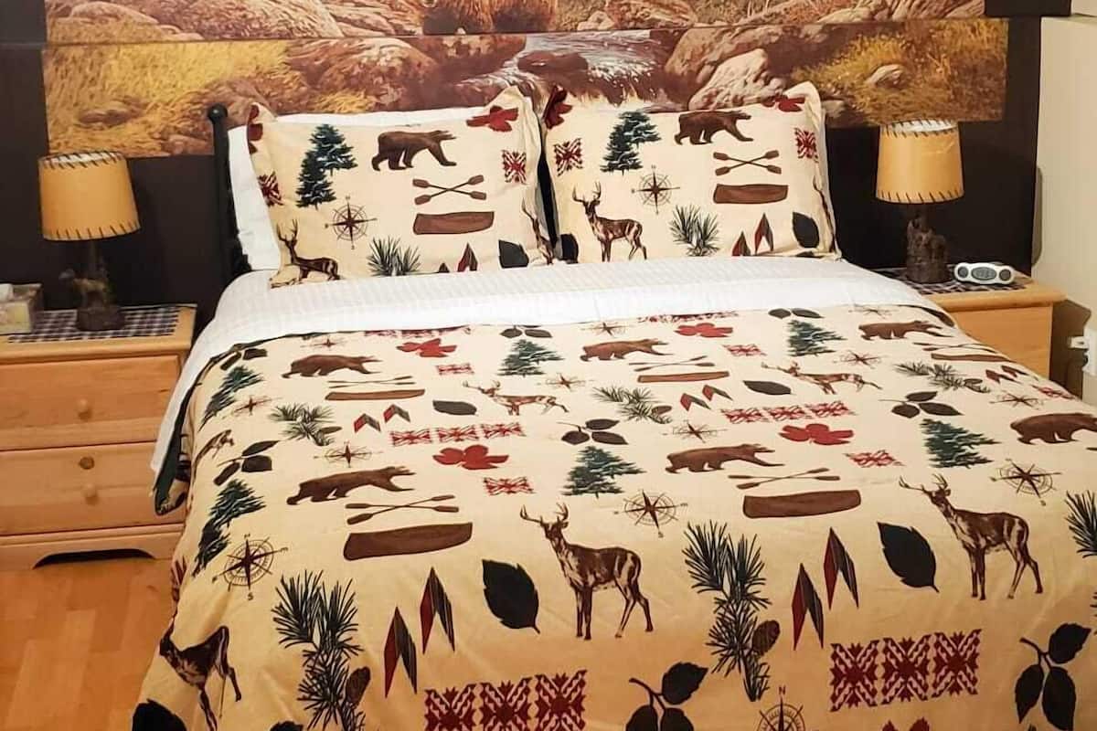 A bed with a forest-themed bedspread, a nightstand with a lamp, and a wall tapestry of a mountainous landscape.