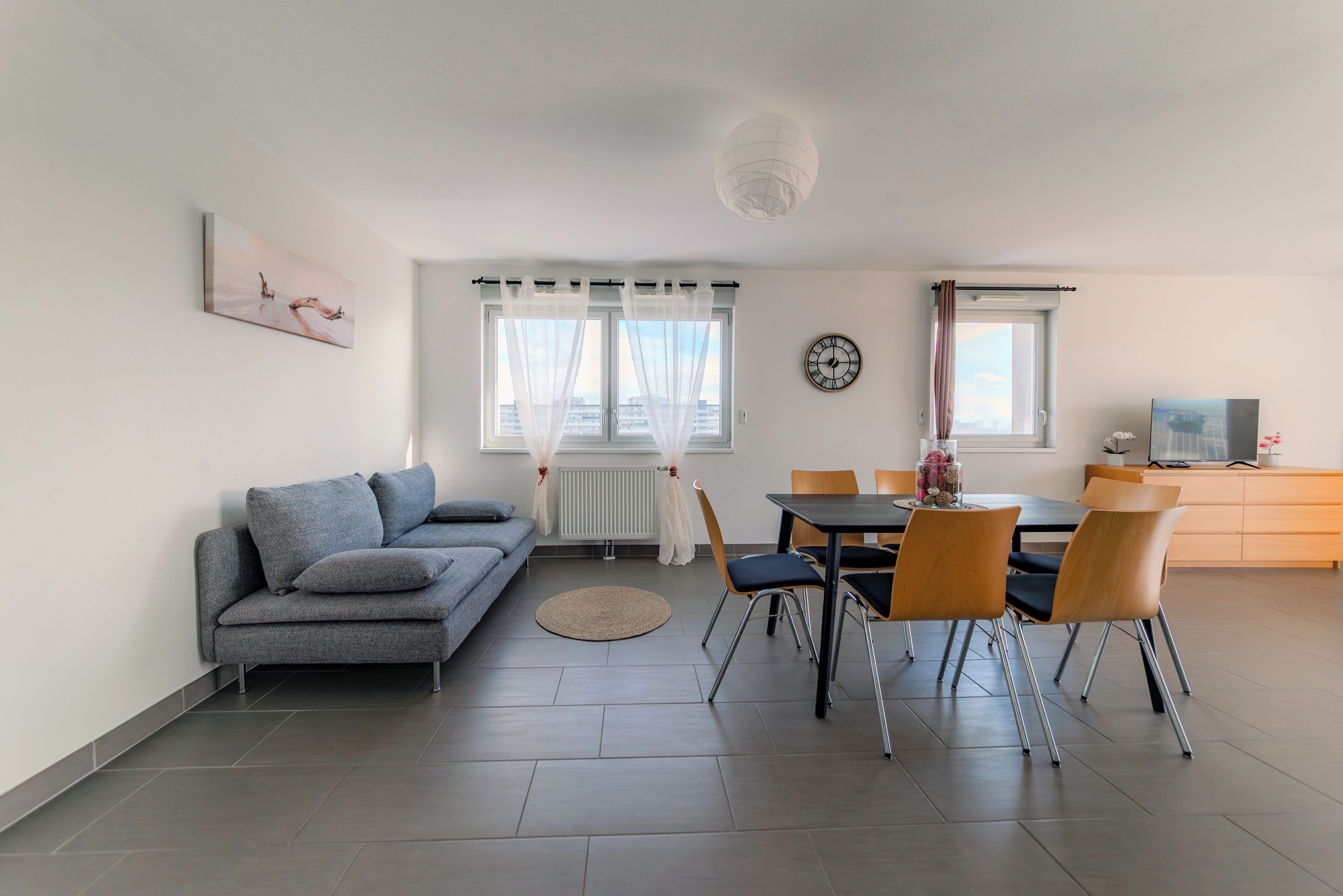 Una sala moderna con un sofá gris, una mesa de comedor con sillas, un reloj en la pared y una ventana con cortinas traslúcidas.