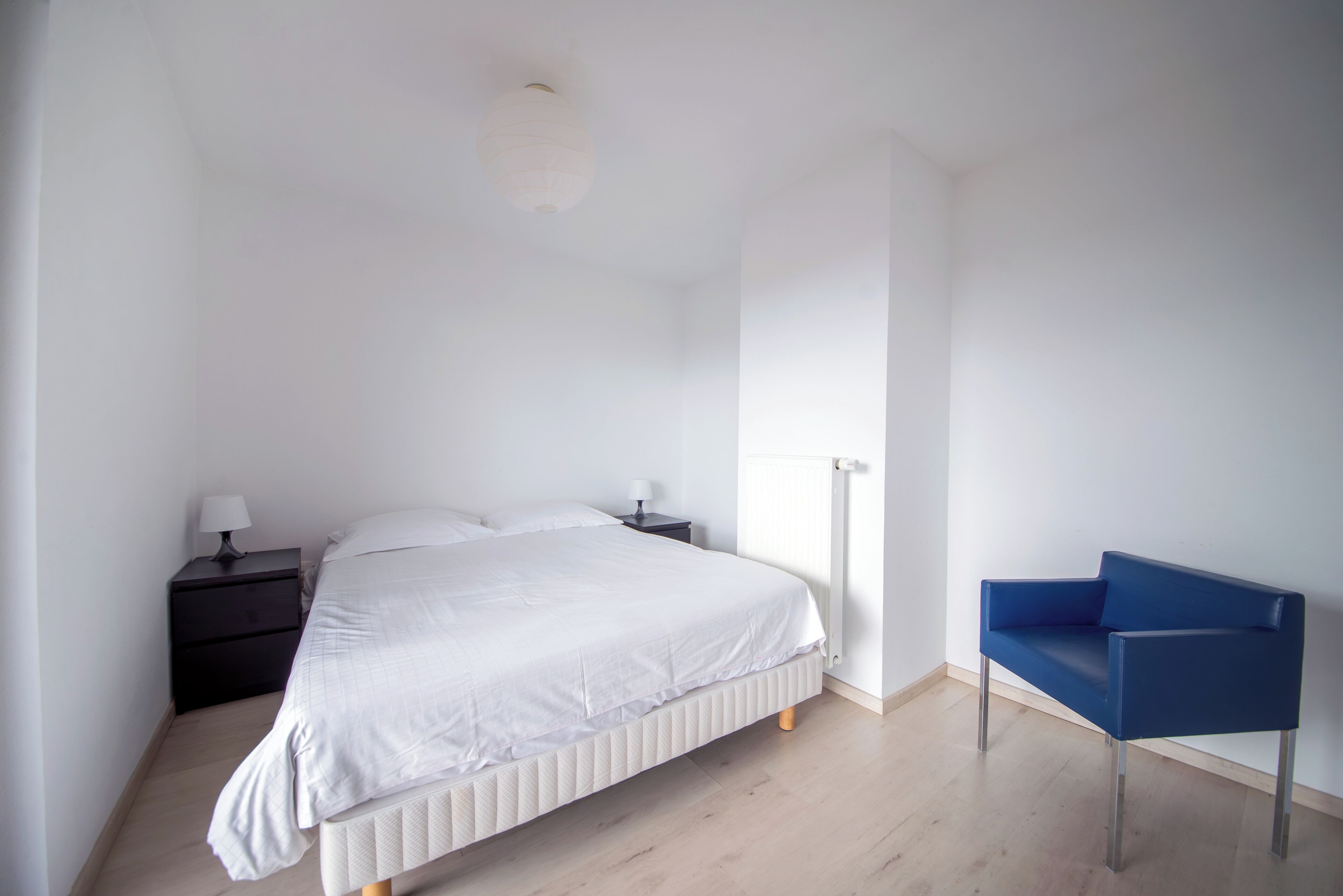 Et soveværelse med en seng, et natbord, en lampe, en radiator og en blå stol.