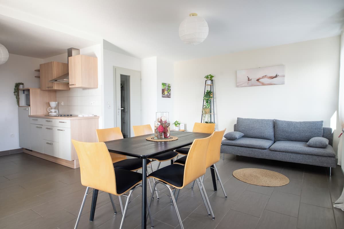 Una cocina moderna con armarios blancos, una mesa de comedor con sillas, un sofá gris y un cuadro en la pared.