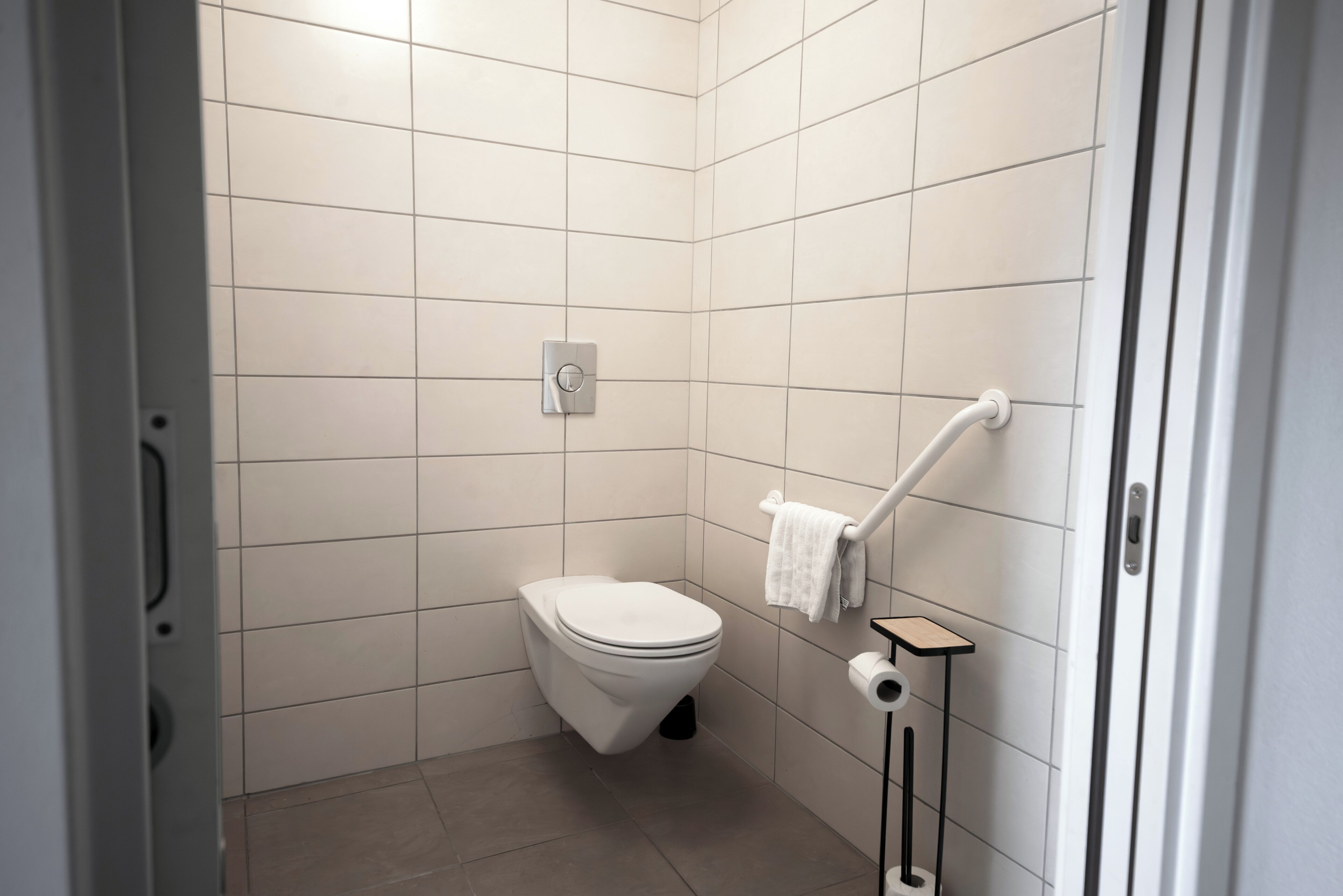 Ein kleines, modernes Badezimmer mit weißen Fliesen, einer Toilette, einem Handtuchhalter und einem Toilettenpapierrollenhalter.