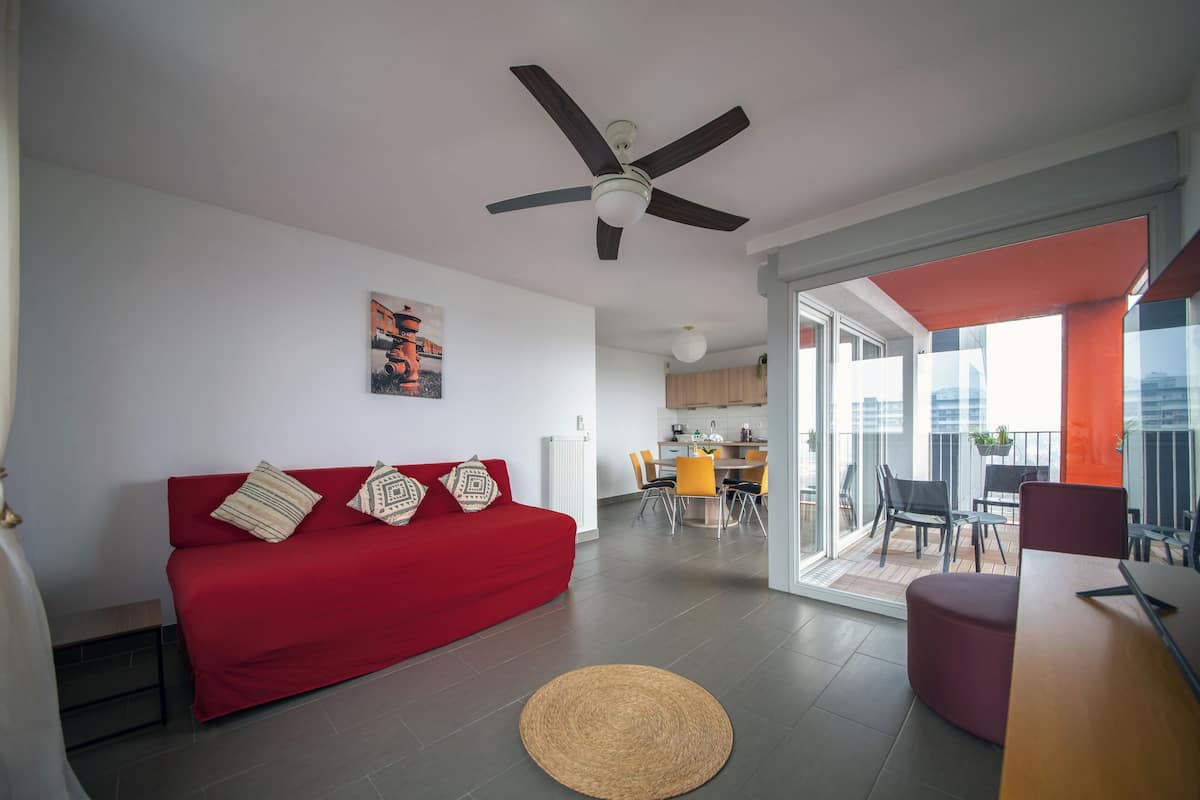 Un moderno salón con un sofá rojo, un ventilador de techo y vistas a un balcón con mobiliario exterior.
