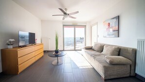 A modern living room with a sofa, TV, wooden cabinet, and a ceiling fan. - Appart du Trident (Mulhouse)