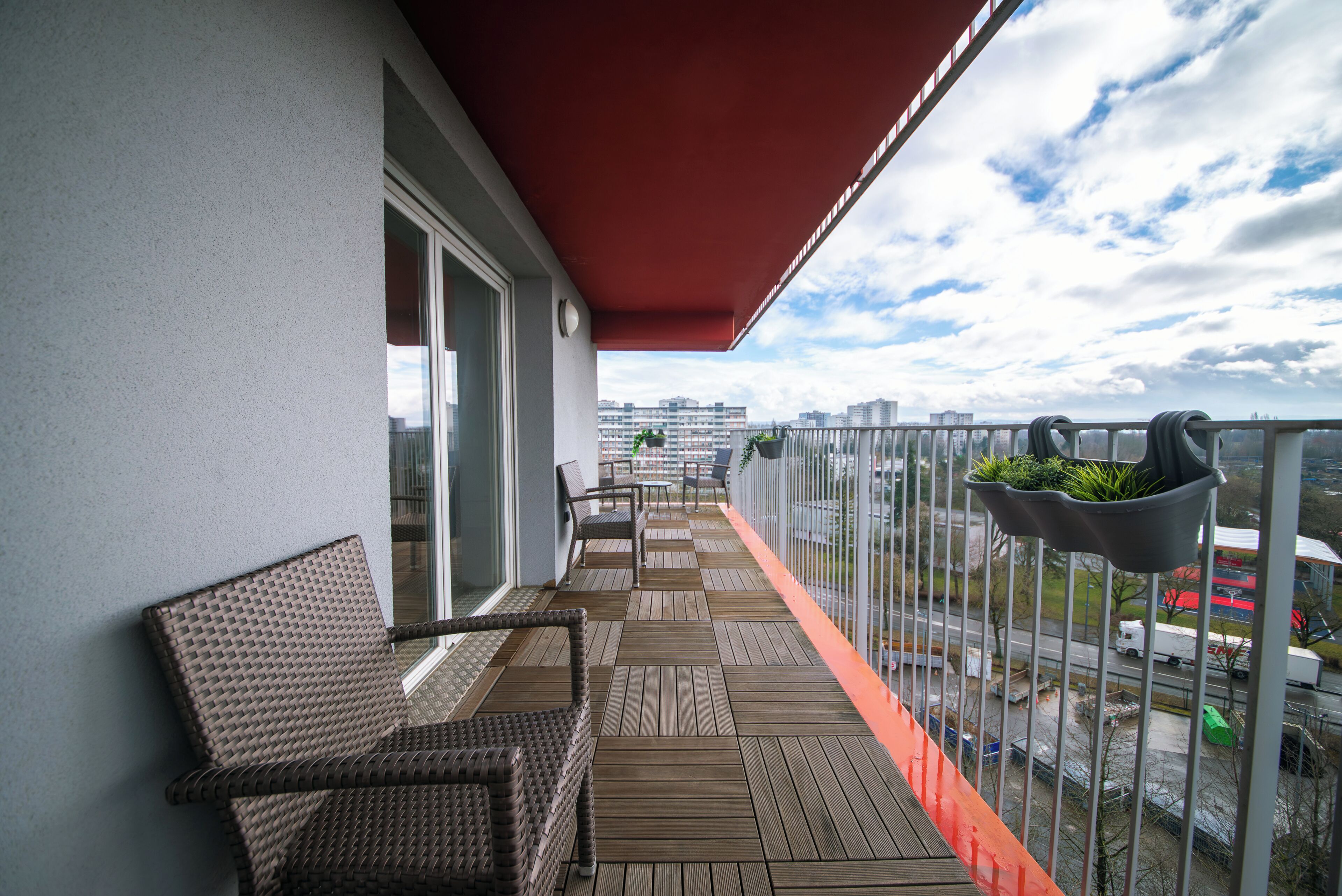 Ein Balkon mit Korbmöbeln, einem roten Markisenstoff und Blick auf die Stadtlandschaft.