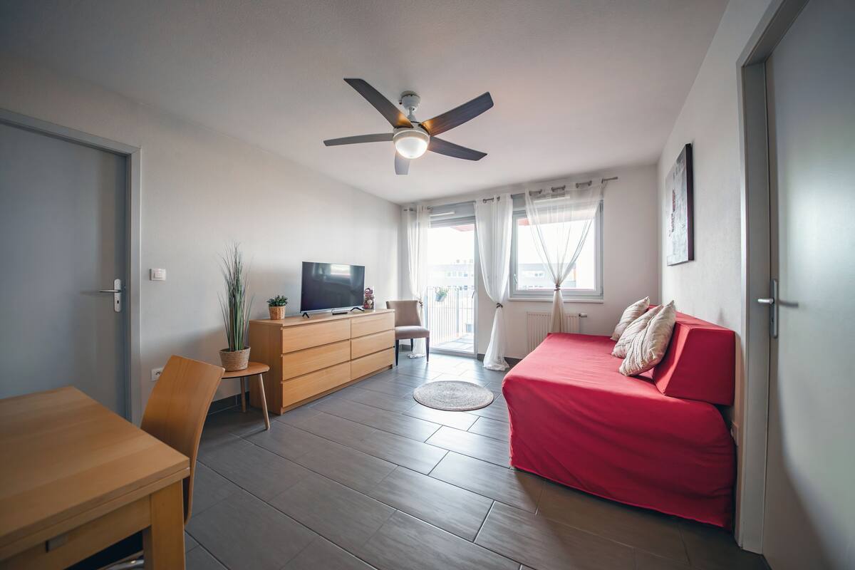 Una habitación con ventilador de techo, un sofá rojo, una mesa de madera, un televisor sobre un mueble de madera y una ventana con cortinas.