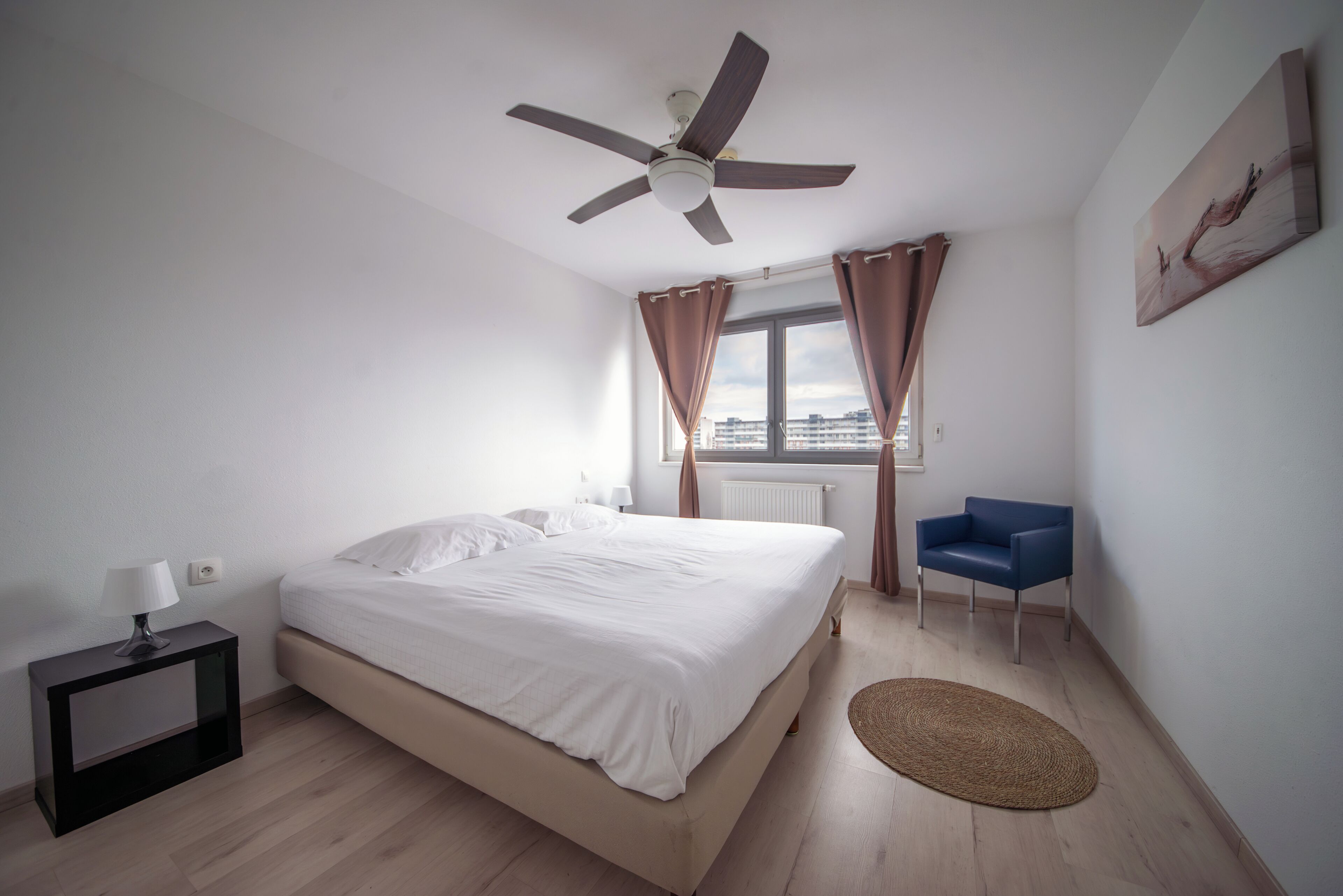 Une chambre avec un lit, un ventilateur de plafond, une fenêtre avec des rideaux, une chaise et une vue sur la ville.