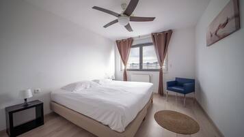 Une chambre avec un lit, un ventilateur de plafond, une fenêtre avec des rideaux, une chaise et une vue sur la ville.