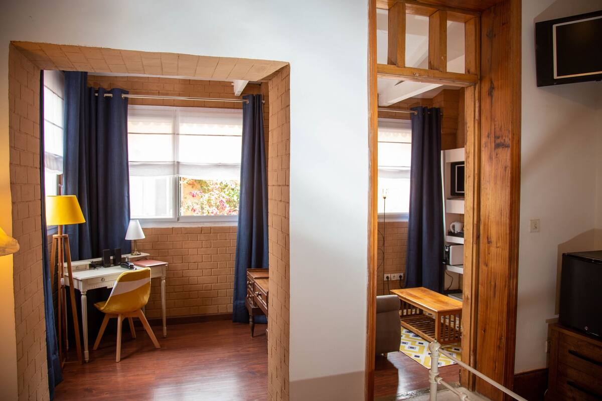 Ein Zimmer mit Schreibtisch, Stuhl und Fernseher. Es gibt ein Fenster mit Vorhängen, eine Lampe und eine Ziegelwand.