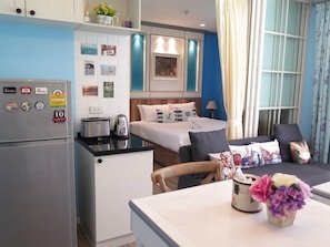 A compact hotel room with a kitchenette, a bed with multiple pillows, a dining table with a vase of flowers, and a window with curtains.