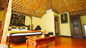 A room with a thatched roof, a bed with a white and green bedspread, a small table with a vase, and a painting on the wall. - Okawati Hotel (Ubud)