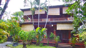 A traditional two-story building with a balcony, surrounded by lush greenery and a stone pathway. - Okawati Hotel (Ubud)