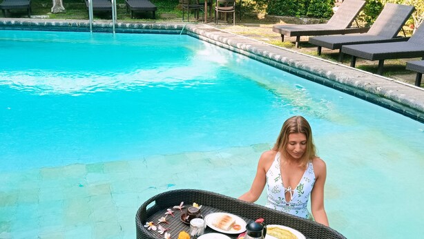 A poolside setting with a wicker table holding food and drinks, surrounded by lounge chairs. - Okawati Hotel (Ubud)