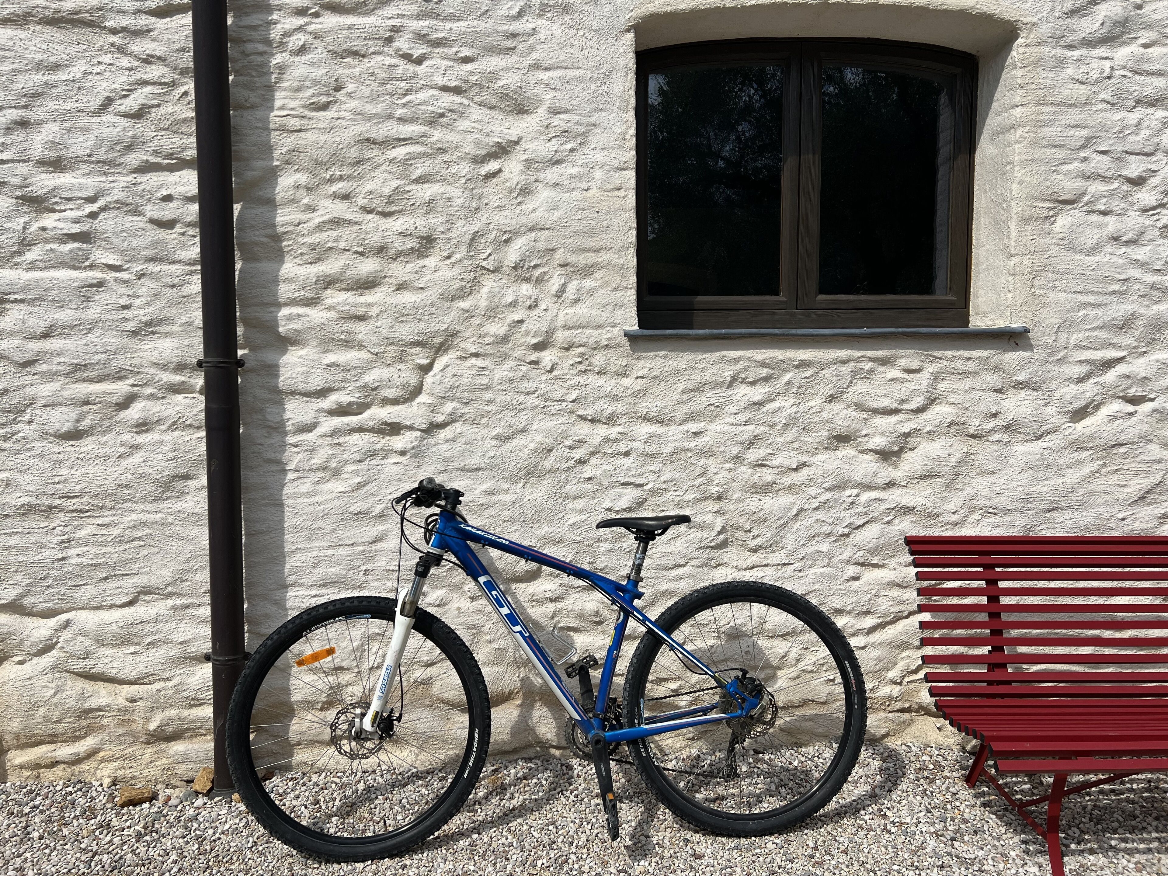 A blue bicycle leaning against a textured wall with a window and a red bench nearby.