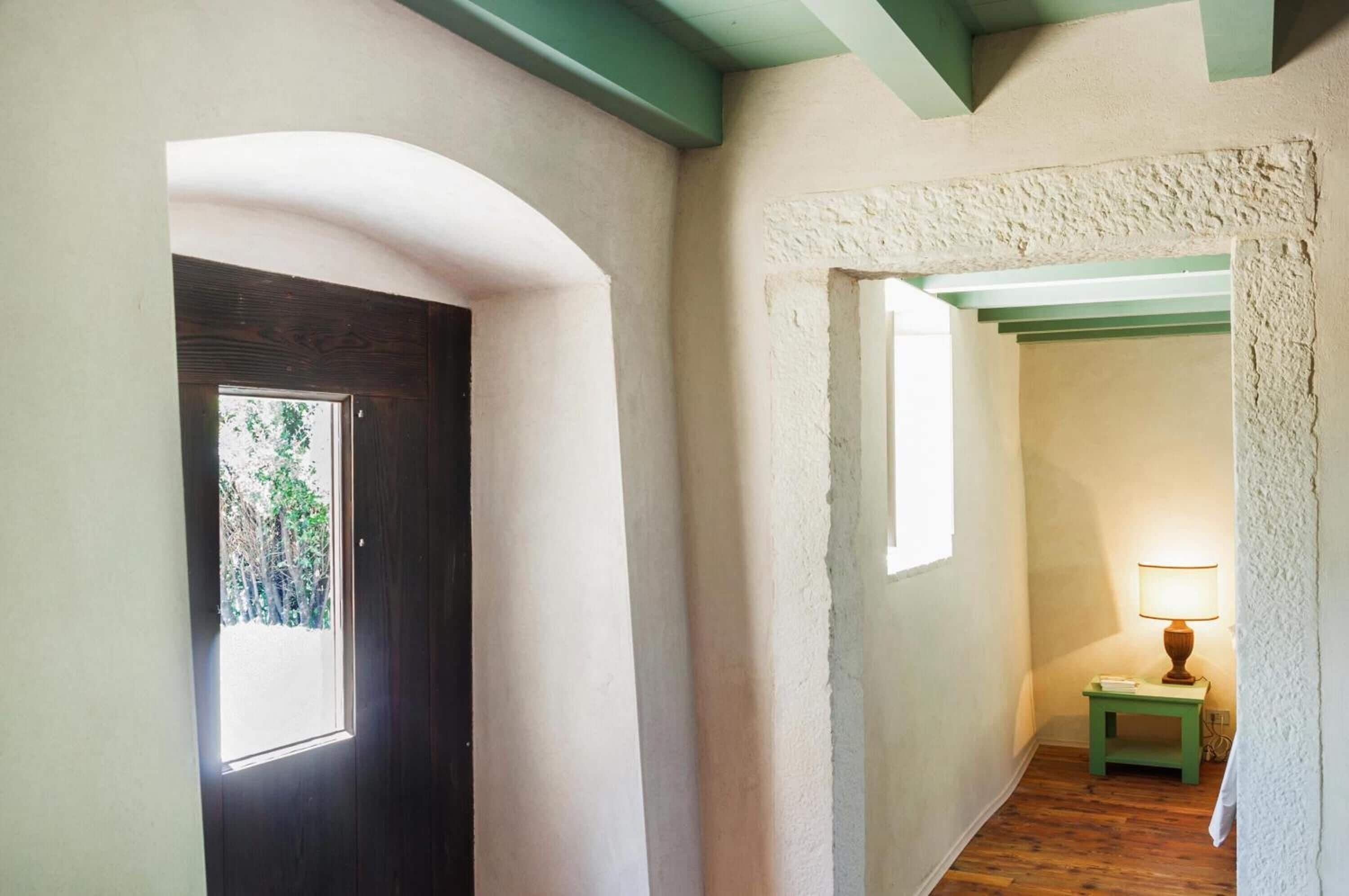 Un pasillo con una puerta de madera, una ventana pequeña y una lámpara sobre una mesita de noche.