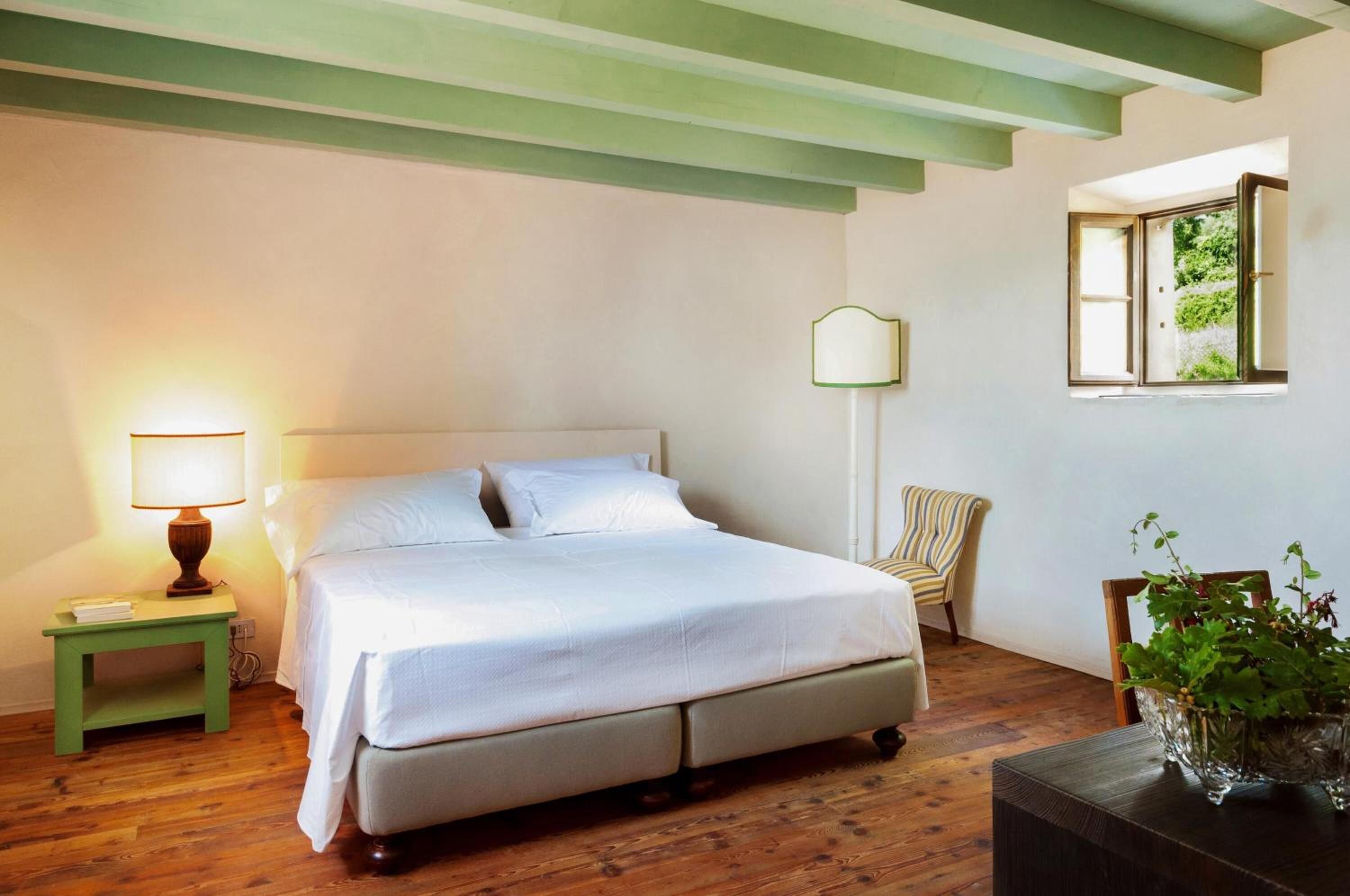 A bedroom with a bed, a nightstand with a lamp, a chair, a window, and a plant.