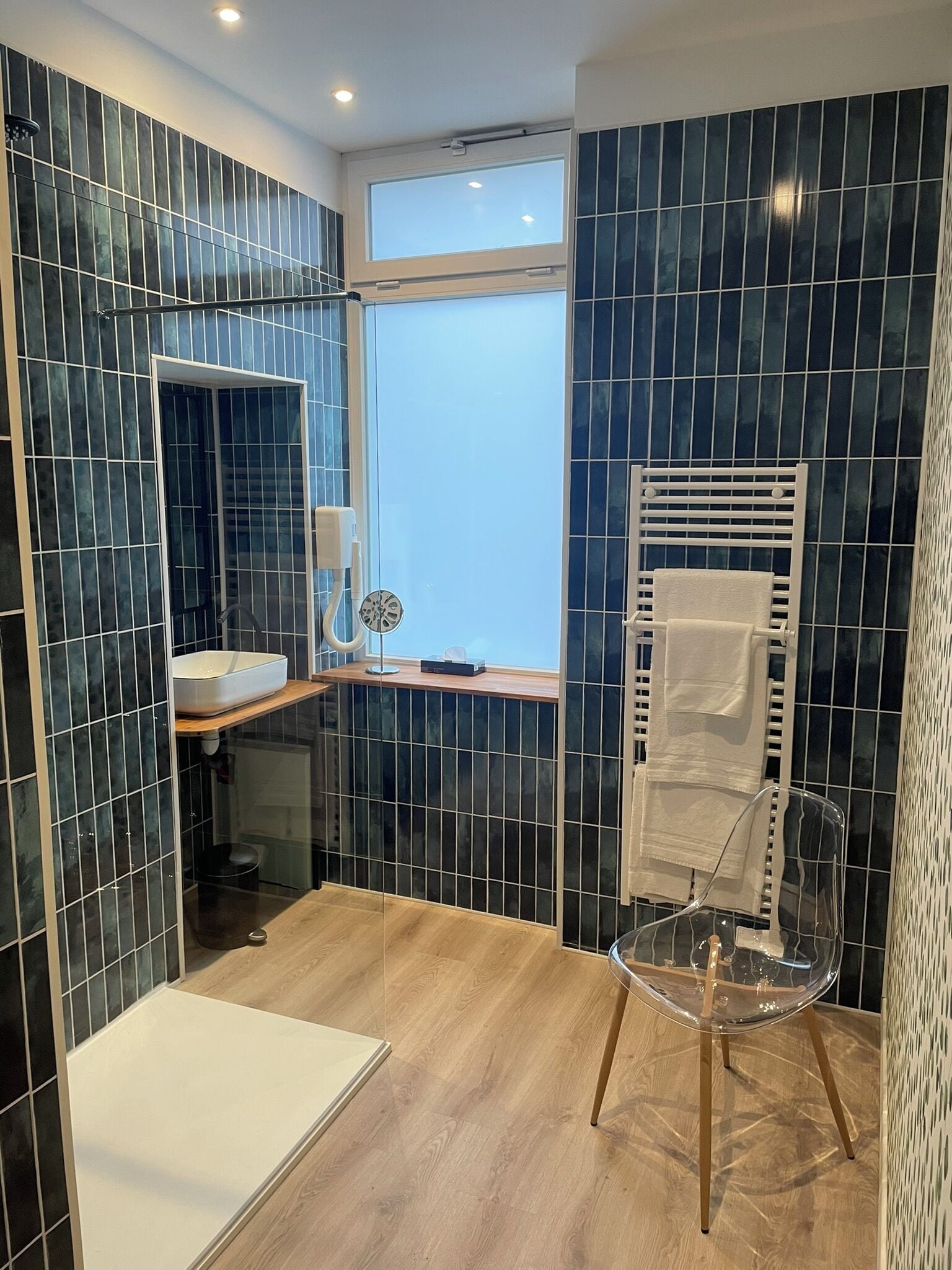 Un baño moderno con azulejos azul oscuro, un lavabo blanco, cabina de ducha con mampara de vidrio y una silla transparente.