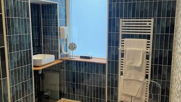 Un baño moderno con azulejos azul oscuro, un lavabo blanco, cabina de ducha con mampara de vidrio y una silla transparente.
