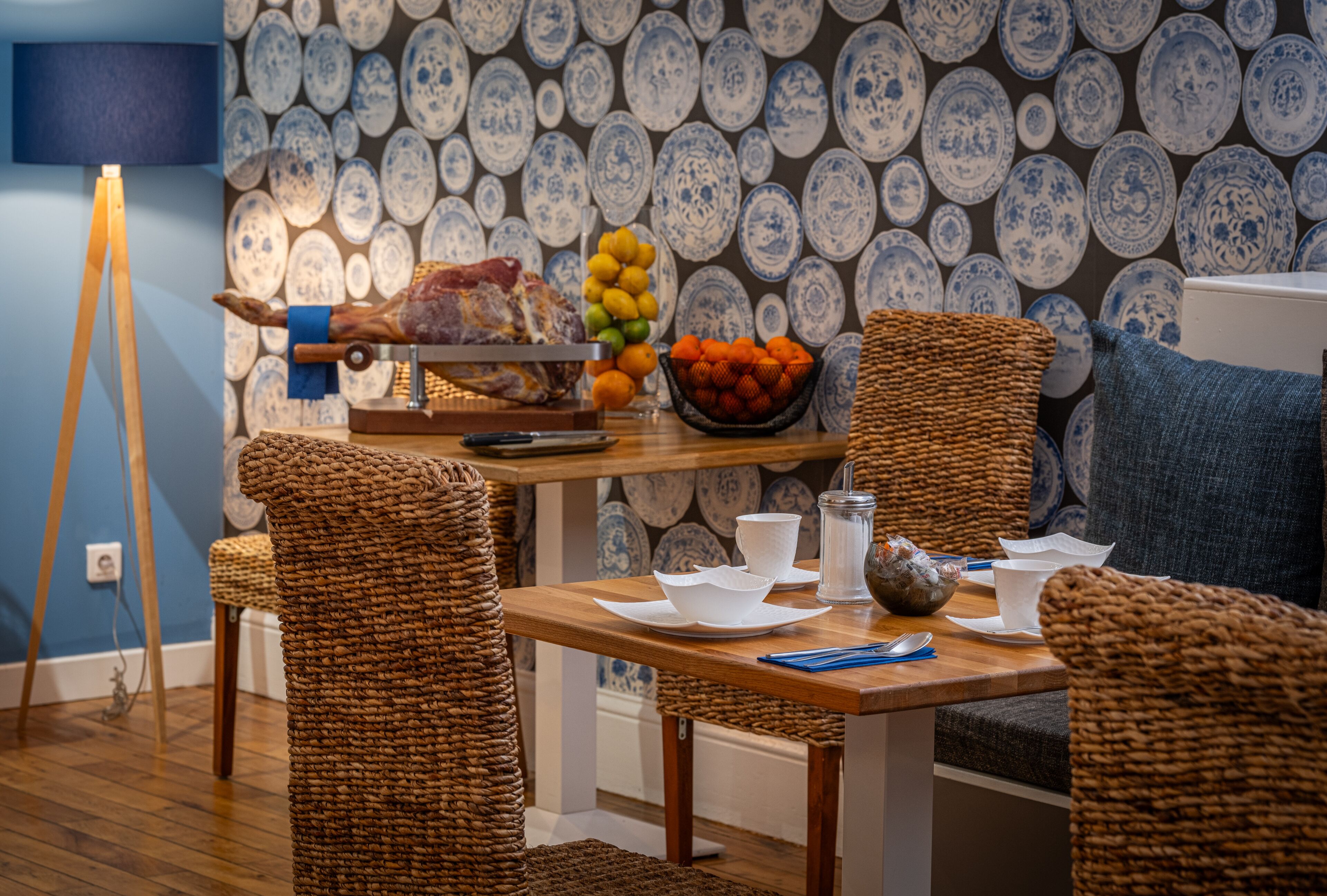 A dining area with a wooden table, wicker chairs, a fruit bowl, and a wall decorated with blue and white plates.