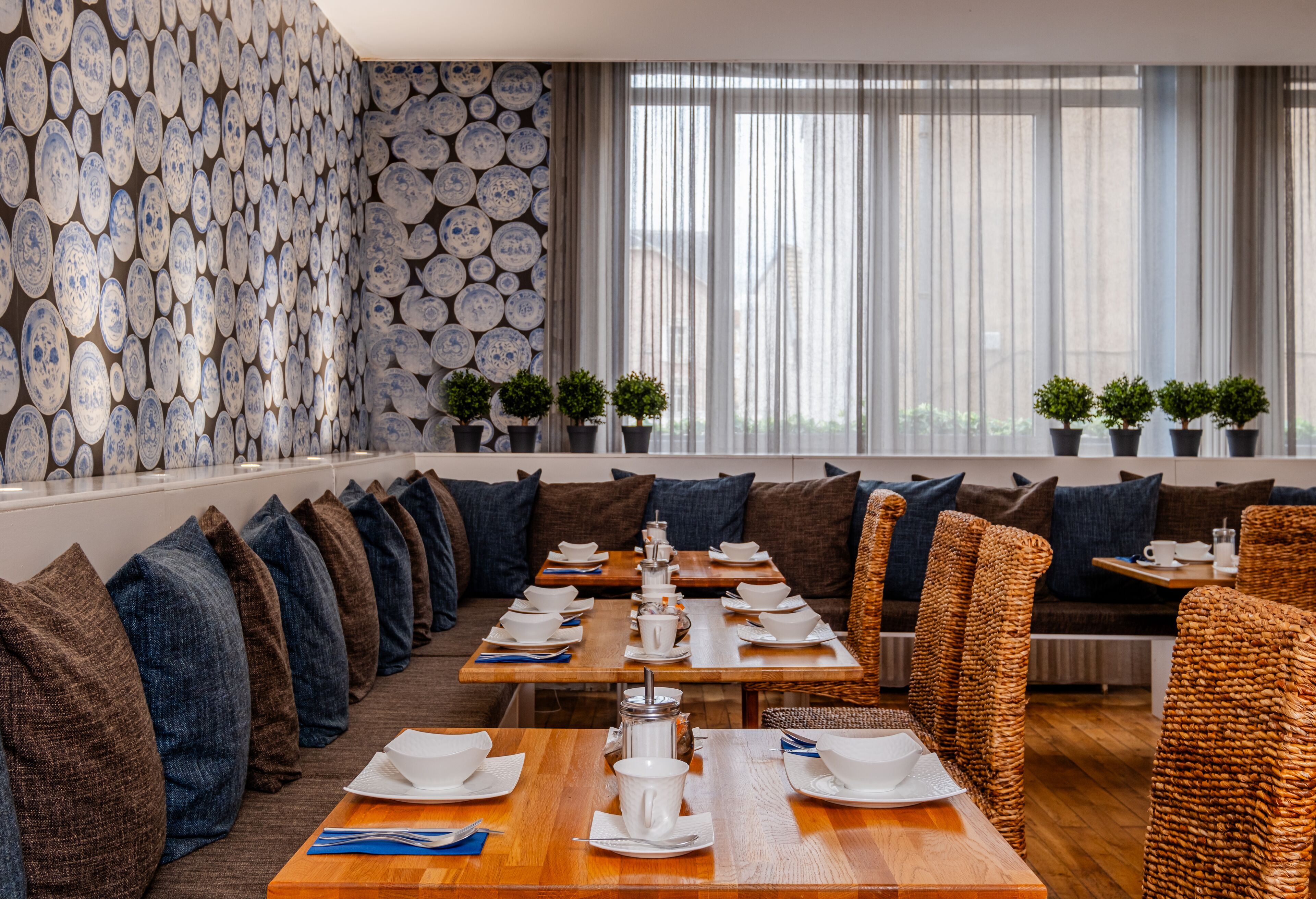A modern dining area with a long table set for a meal, surrounded by sofas and chairs, with a decorative wall of plates and potted plants.