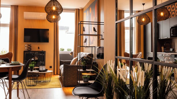 A modern living room with a sofa, dining table, and chairs, a TV, and a kitchen area visible through a glass partition. - Nouvel Hôtel & Appartements (Oyonnax)
