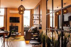 A modern living room with a sofa, dining table, and chairs, a TV, and a kitchen area visible through a glass partition. - Nouvel Hôtel & Appartements (Oyonnax)