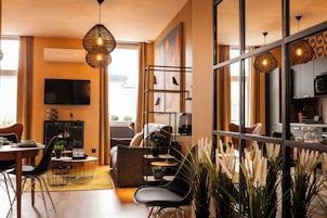 Una sala de estar moderna con sofá, mesa de comedor y sillas, televisor y una cocina visible a través de una pared de vidrio.