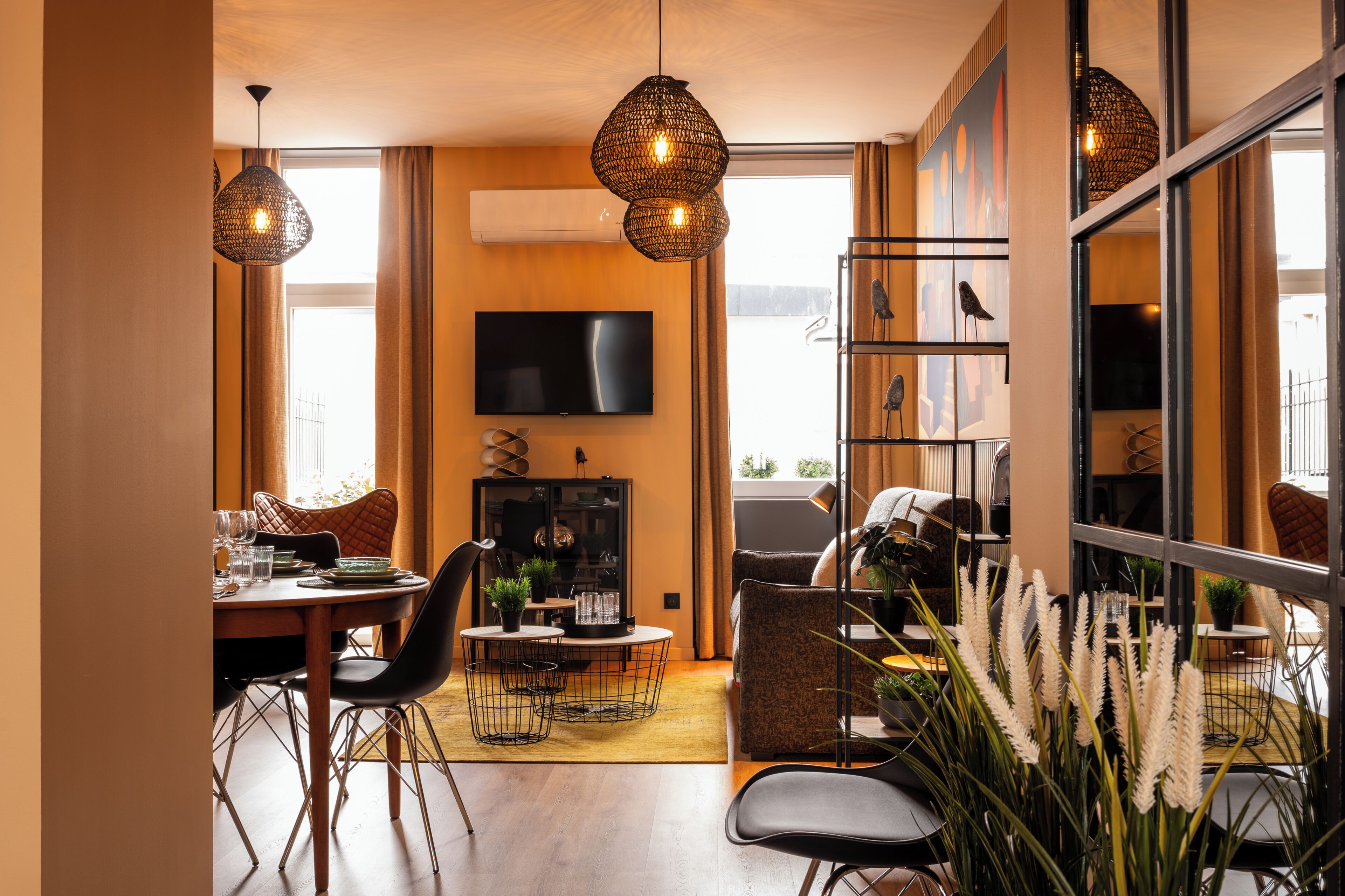 A modern living room with a dining area, a flat-screen TV mounted on the wall, and a large window with curtains.