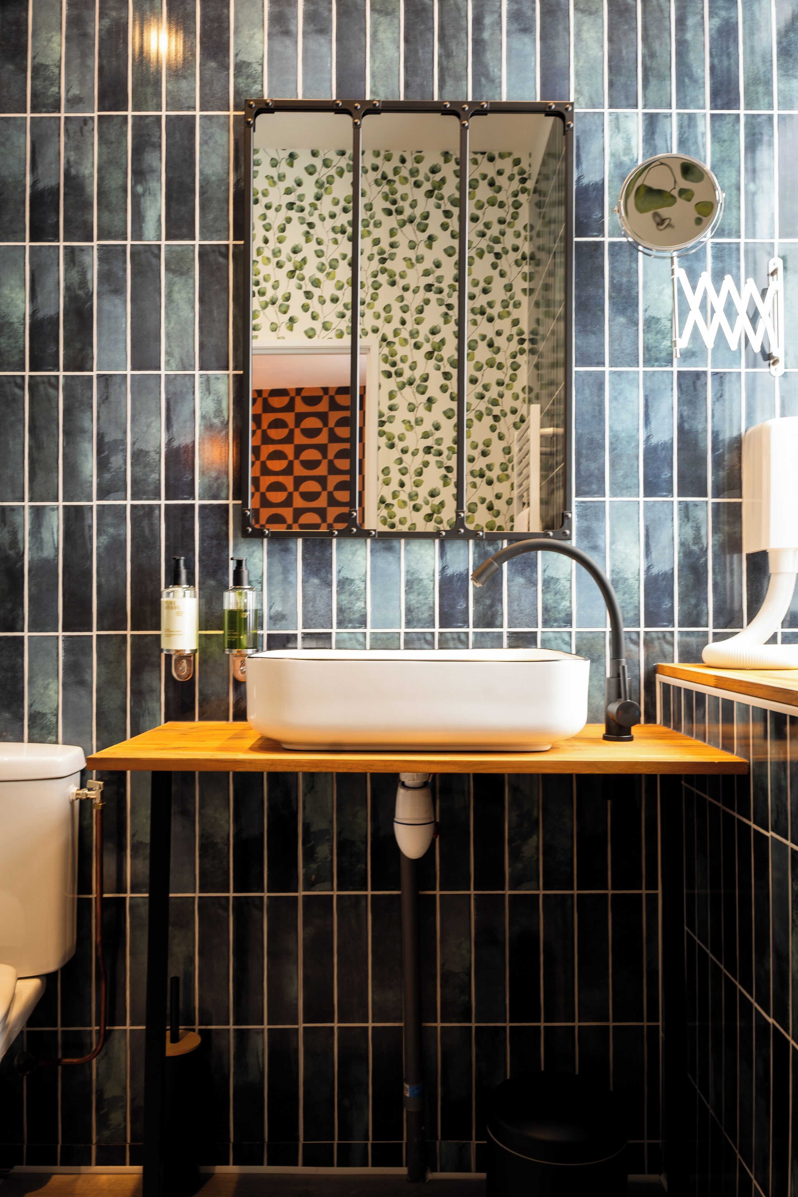 Une salle de bain avec un lavabo blanc, des murs recouverts de carreaux noirs et un miroir orné d’un motif de feuilles.