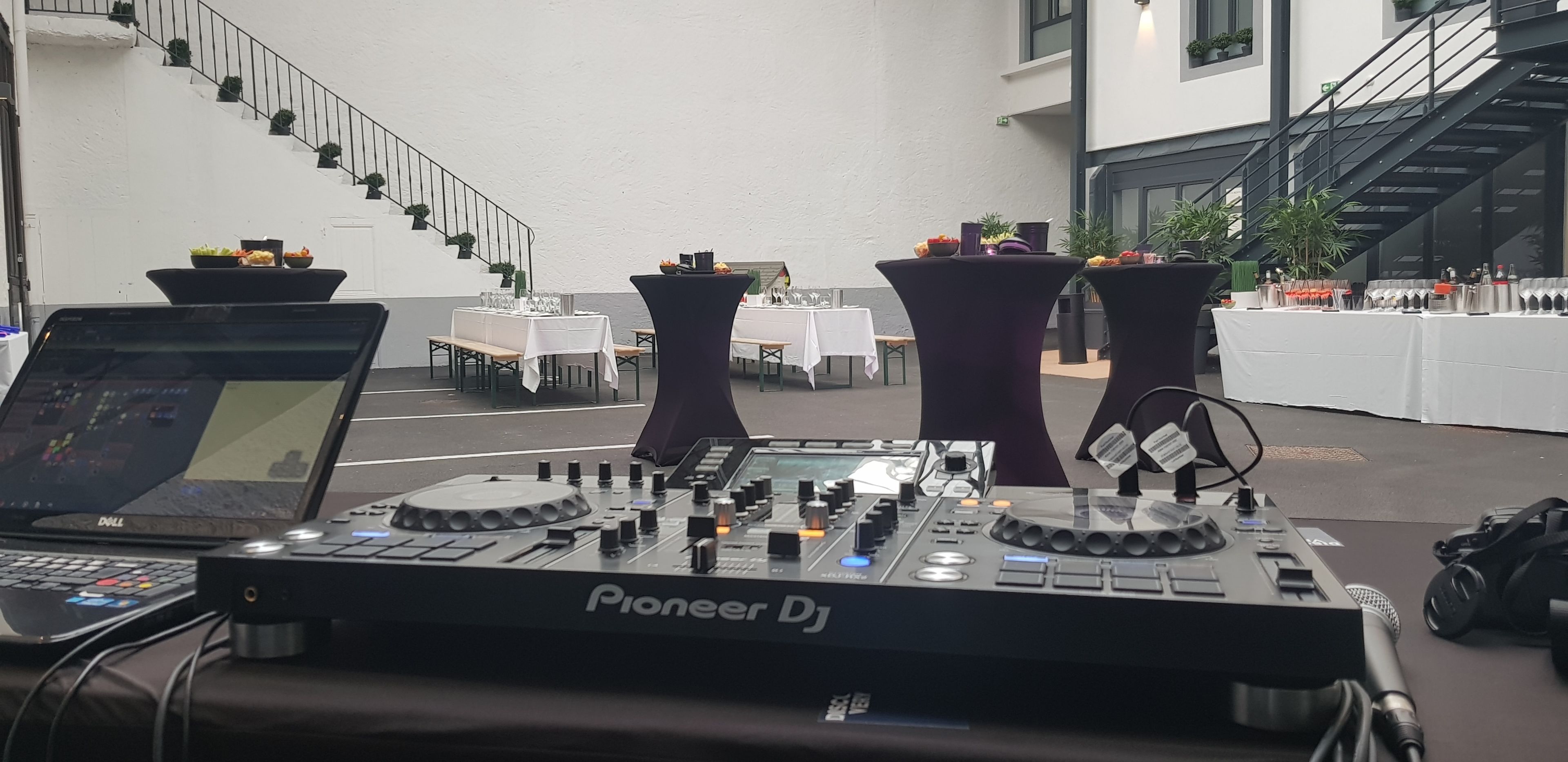 A Pioneer DJ audio mixing console in a modern indoor setting with tables and chairs, a staircase, and a white wall.