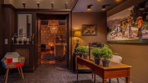 A hallway with a bar area, a seating area with a patterned chair, a wooden table with potted plants, and a painting on the wall. - Nouvel Hôtel & Appartements (Oyonnax)