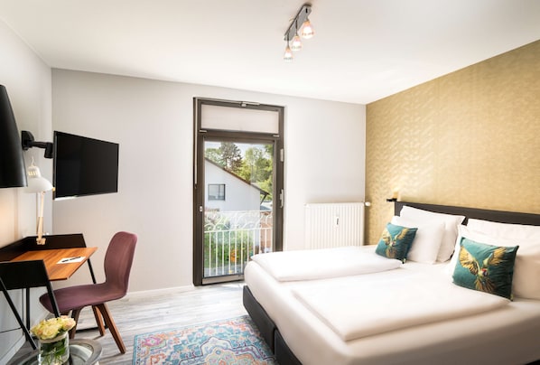 A modern hotel room with a large bed, a desk with a chair, a TV, and a balcony with a view of greenery.