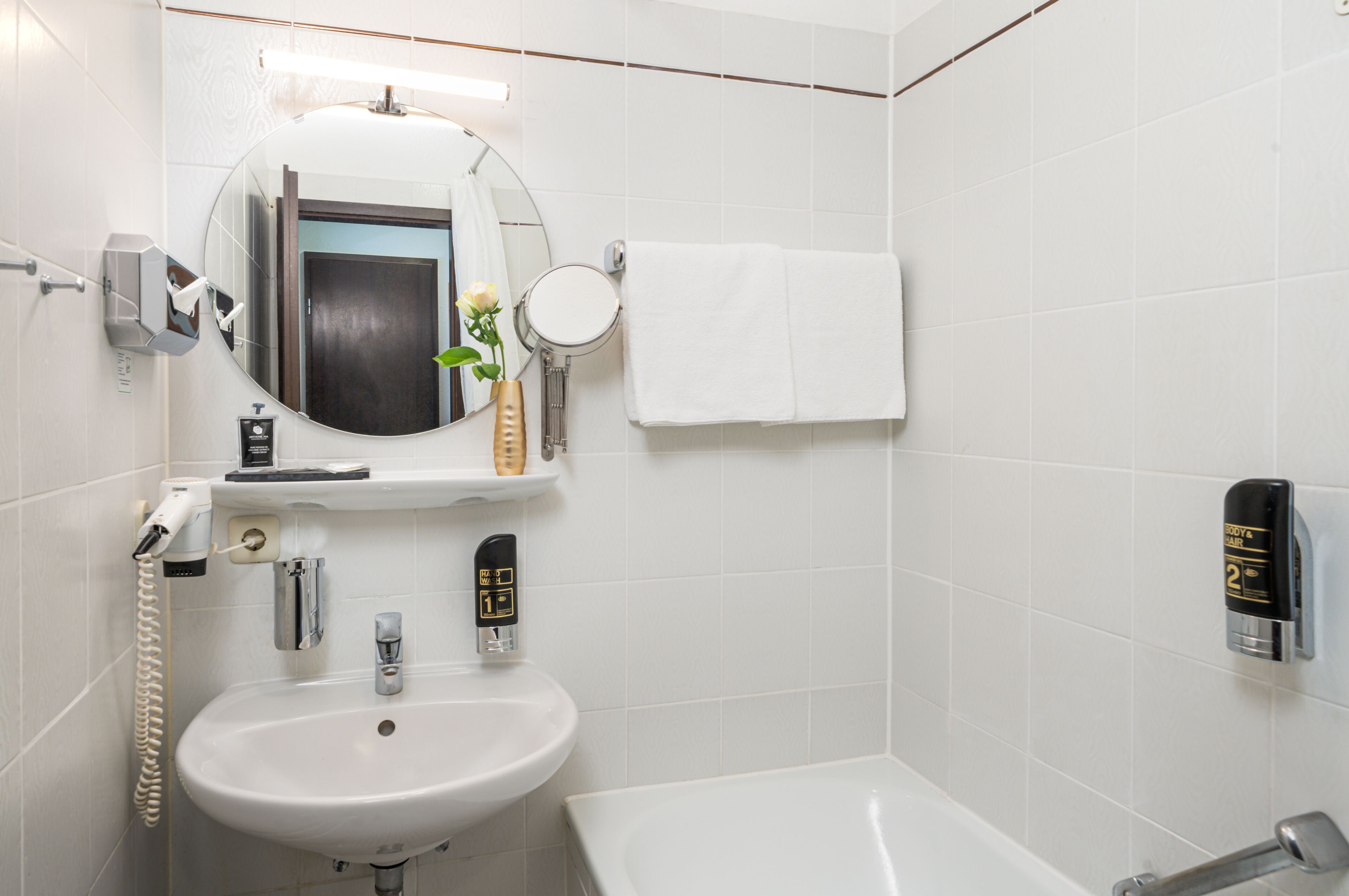 Un baño moderno con un lavamanos blanco, un espejo redondo, un espejo de mano, un perchero para toallas, un dispensador de jabón y un secamanos automático.