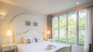 A neatly made bed with white bedding and pillows, two bedside tables with lamps, and a large window with curtains. - Chelona Huahin Condo Garden View by Dome (Hua Hin)